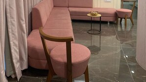 A modern living room with a pink sectional sofa, a small round table, and a wooden chair. - Pam Inn Hotel Vadodara (Vadodara)