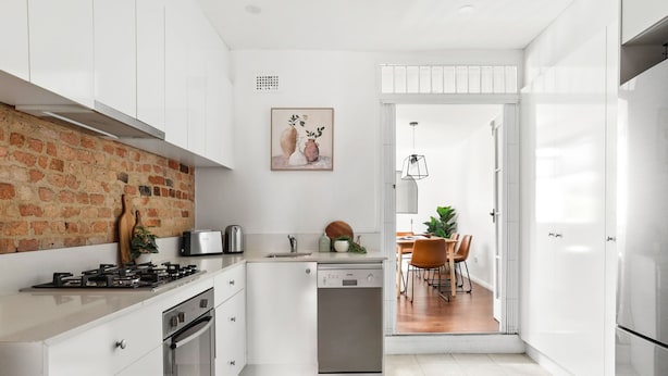 A modern kitchen with white cabinetry, a brick accent wall, a dishwasher, and a dining area with a table and chairs.