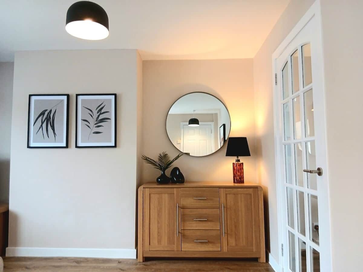 A hallway with a wooden cabinet, a round mirror, and two framed artworks on the wall.