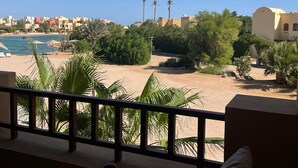 Blick von einem Balkon auf einen Sandstrand mit Palmen, einen Pool und Gebäude im Hintergrund.