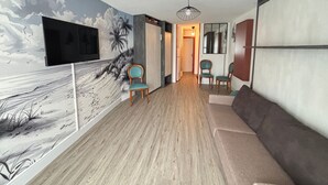 Sala de estar com um mural, piso de madeira, um sofá, duas poltronas, uma televisão e uma luminária no teto.