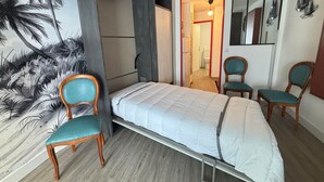 Quarto compacto com uma cama, duas cadeiras, guarda-roupa e uma parede com um mural de palmeiras.