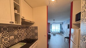 Cozinha com ladrilhos estampados, pia e fogão. Há uma área de jantar com mesa e cadeiras, e uma sala de estar com sofá e televisão.