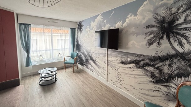 Quarto com um mural de praia, uma mesa redonda, uma cadeira e uma televisão fixada na parede.