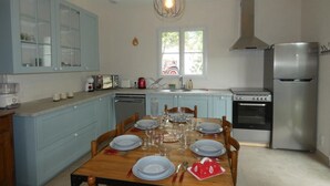 A kitchen with light blue cabinets, a wooden dining table set for four, a stainless steel refrigerator, and a window with a view of greenery outside. - Vendée House 3 BR, Pet-Friendly, Near Beach (Notre-Dame-de-Monts)