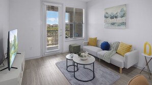 A modern living room with a grey sofa, a small green ottoman, and a round coffee table. There is a painting on the wall and a television on the left. - Charming 1-bedroom Haven I Frisco Dallas I Pool / Gym / Work Space (Frisco)