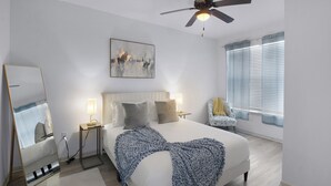 A bedroom with a bed, bedside lamps, a ceiling fan, a window with blinds, a chair, and a painting on the wall. - Charming 1-bedroom Haven I Frisco Dallas I Pool / Gym / Work Space (Frisco)