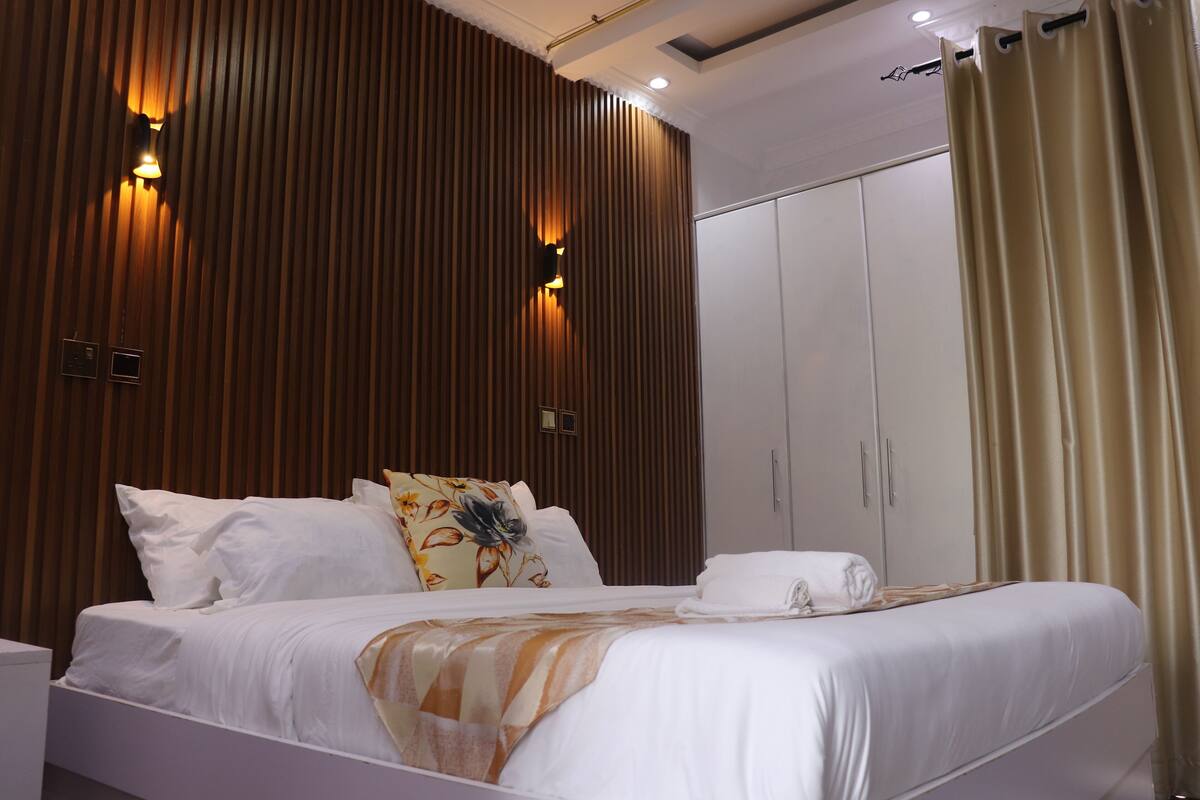 A modern hotel room with a large bed, wooden paneling on the walls, a wardrobe, and a curtain.