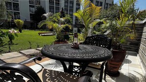 Une terrasse extérieure avec une table ronde et des chaises, entourée de plantes en pot et d’une pelouse.
