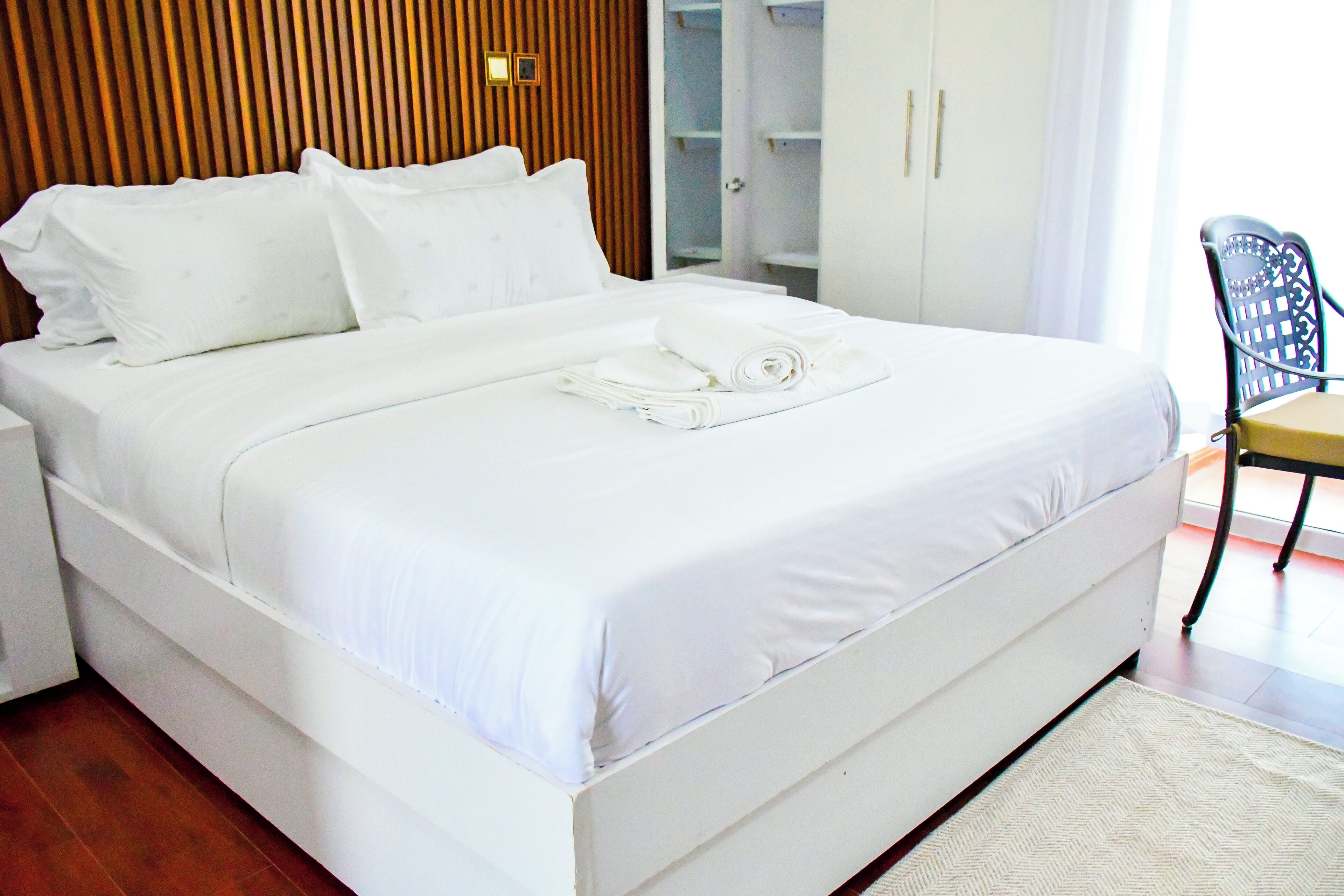 Una cama bien tendida con sábanas blancas y una toalla enrollada, un cabecero de madera, un armario con las puertas abiertas y una silla con cojín.