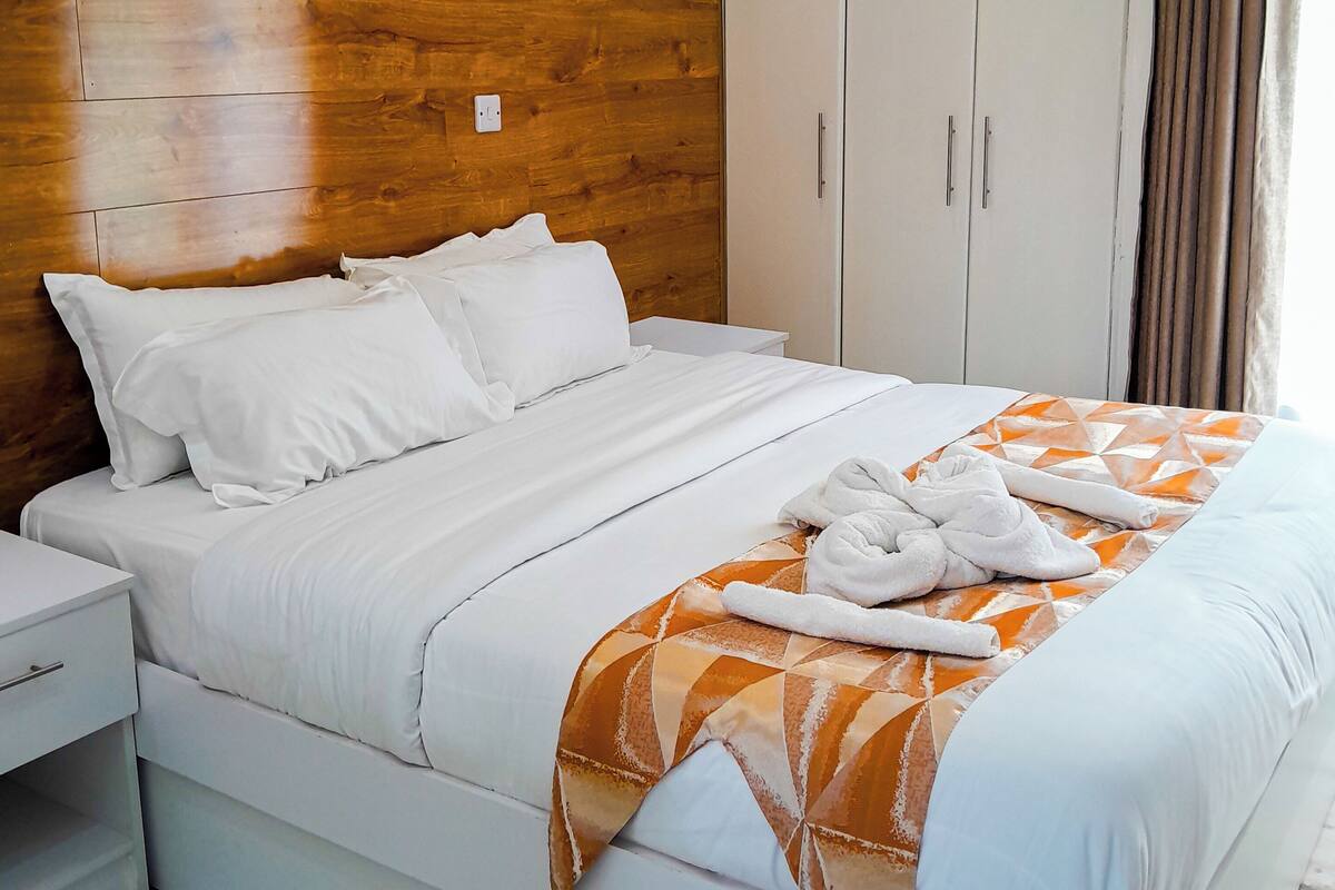 A hotel room with a wooden headboard, a bed with white bedding, a patterned throw blanket, and a wardrobe.