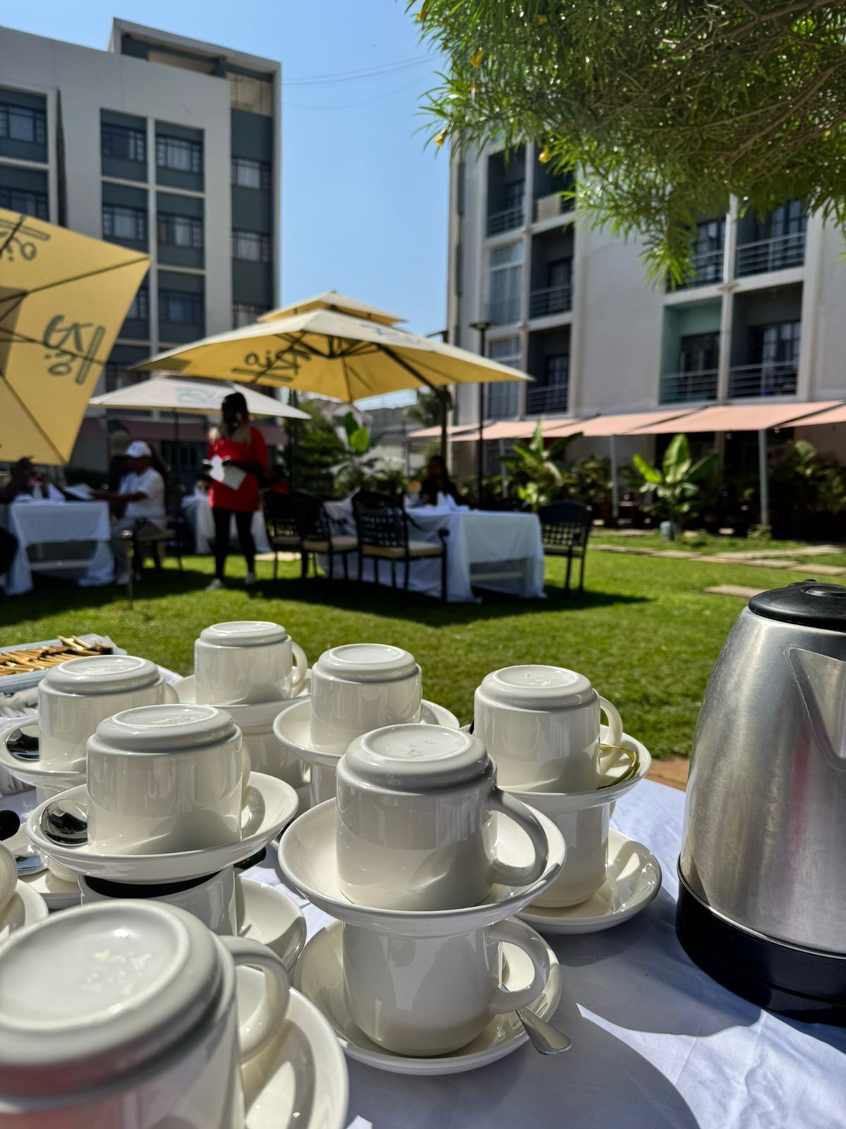 A table set with white cups and saucers, a silver teapot, and a yellow umbrella in an outdoor dining area with tables and chairs, and a building in the background.