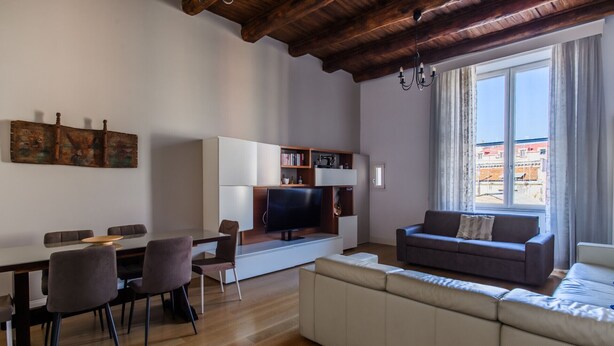 A modern living room with a wooden ceiling, a dining area with a table and chairs, a sofa, and a TV on a white cabinet.