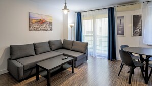 Ένα μοντέρνο σαλόνι με έναν γκρι γωνιακό καναπέ, ένα μικρό τραπεζάκι και μια τραπεζαρία με ένα στρογγυλό τραπέζι και καρέκλες. Στον τοίχο υπάρχει ένας πίνακας, ένα παράθυρο με κουρτίνες και μια μονάδα κλιματισμού.