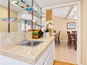 A kitchen with a granite countertop, stainless steel sink, and a glass shelf with decorative items.