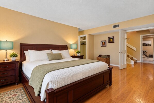 A bedroom with a wooden bed, bedside tables, a nightstand, a lamp, a plant, and a view of a staircase leading to another room.