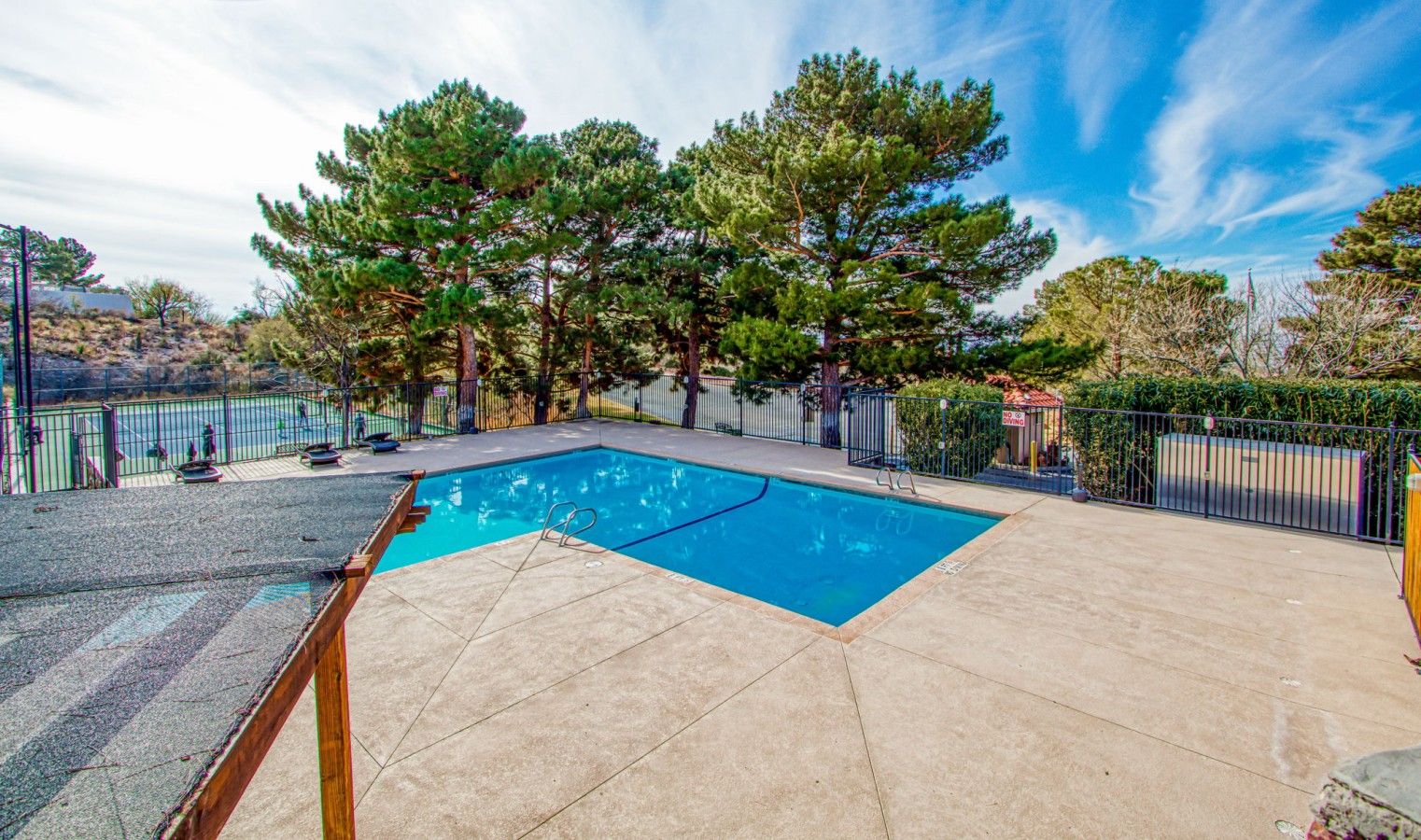 A rectangular swimming pool with a wooden deck and metal railings, surrounded by trees and a fenced area.