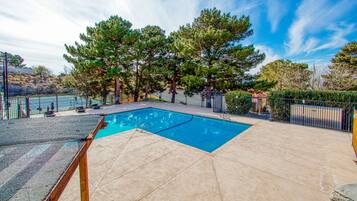 A rectangular swimming pool with a wooden deck and metal railings, surrounded by trees and a fenced area.
