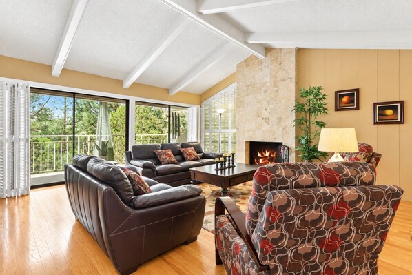 A spacious living room with a fireplace, a balcony with a view of trees, and a large window.