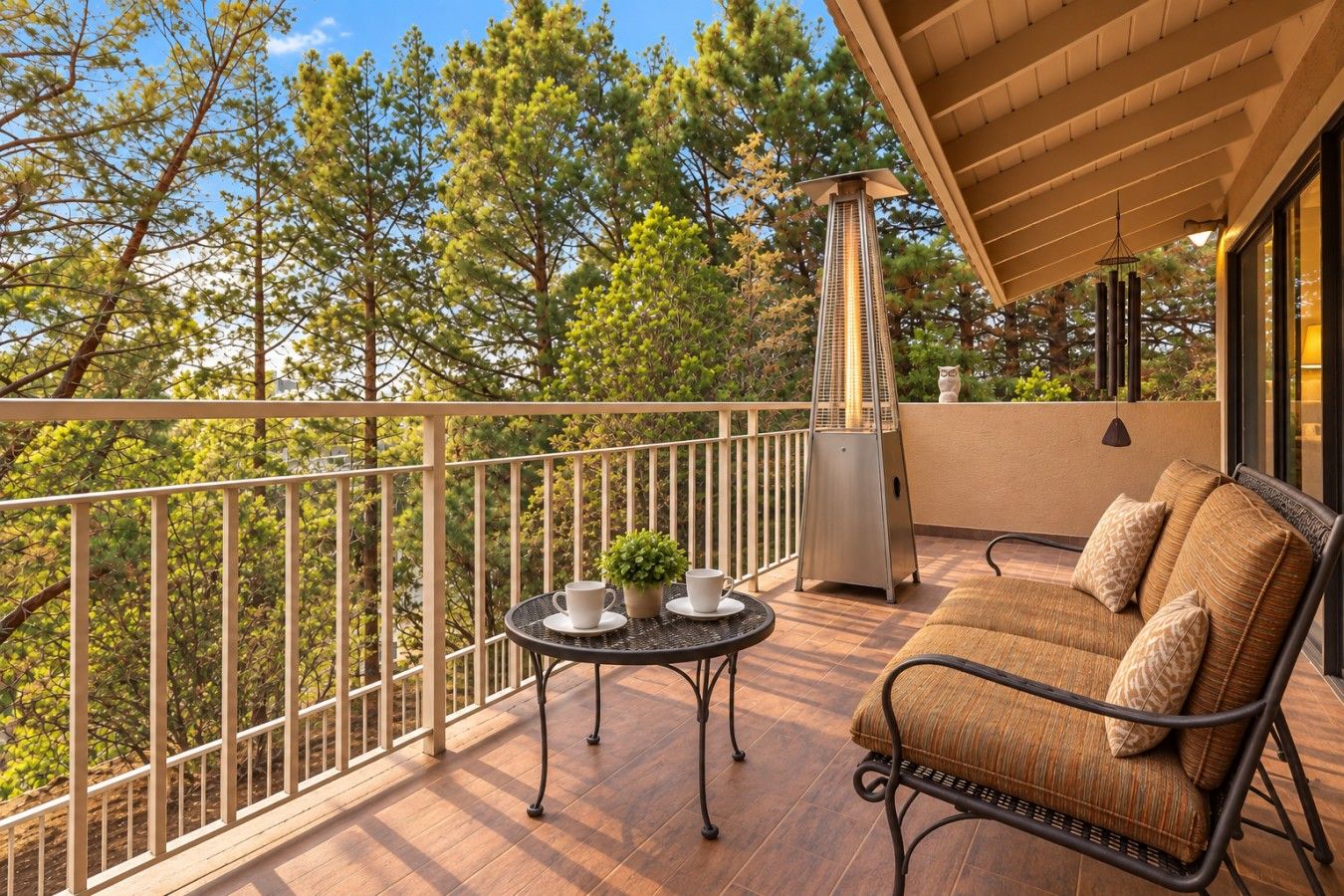 A balcony with a wooden floor, a round table with a cup and a plant, a sofa with cushions, and a metal heater.