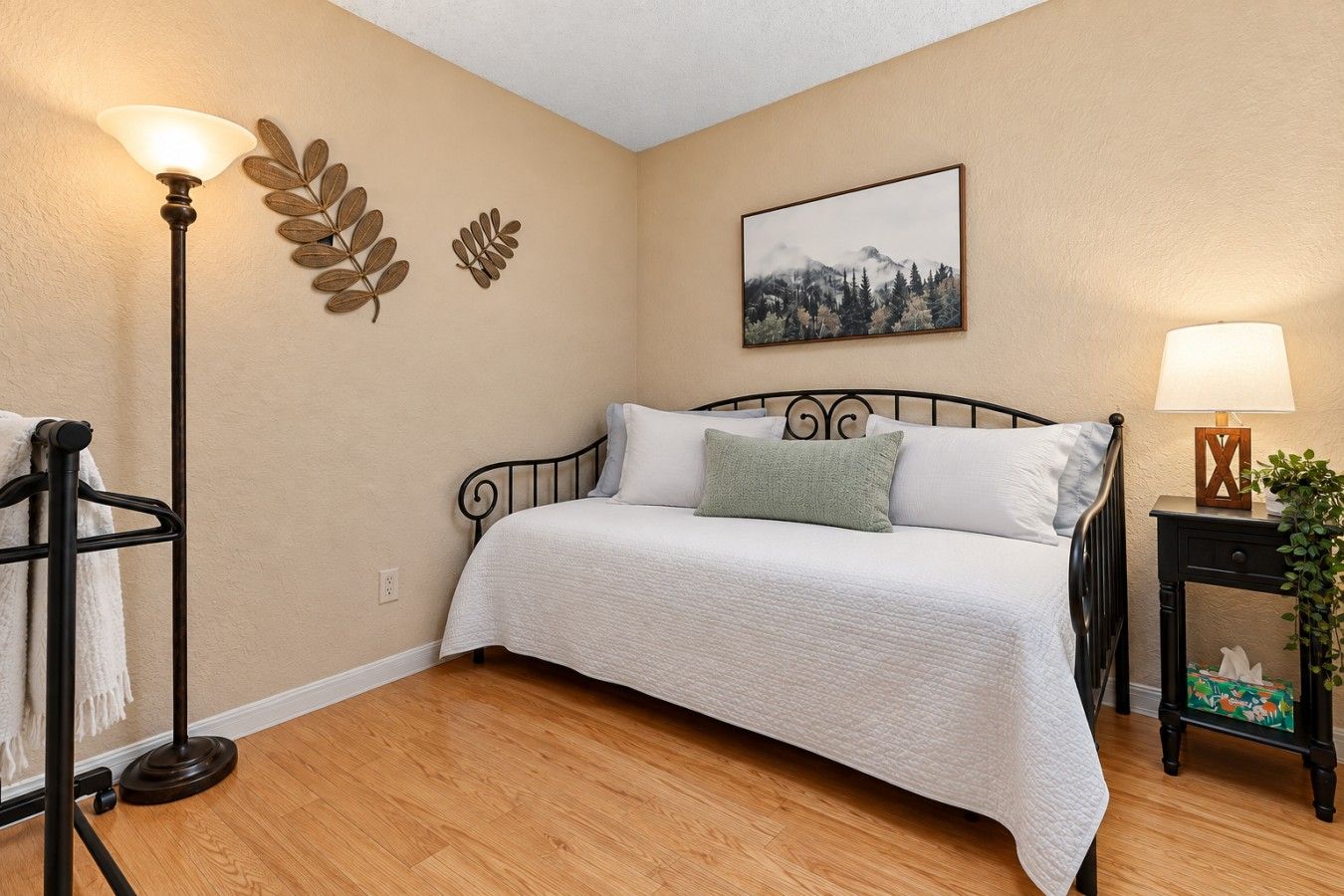A bedroom with a bed, a nightstand with a lamp, a wall-mounted plant, and a painting.