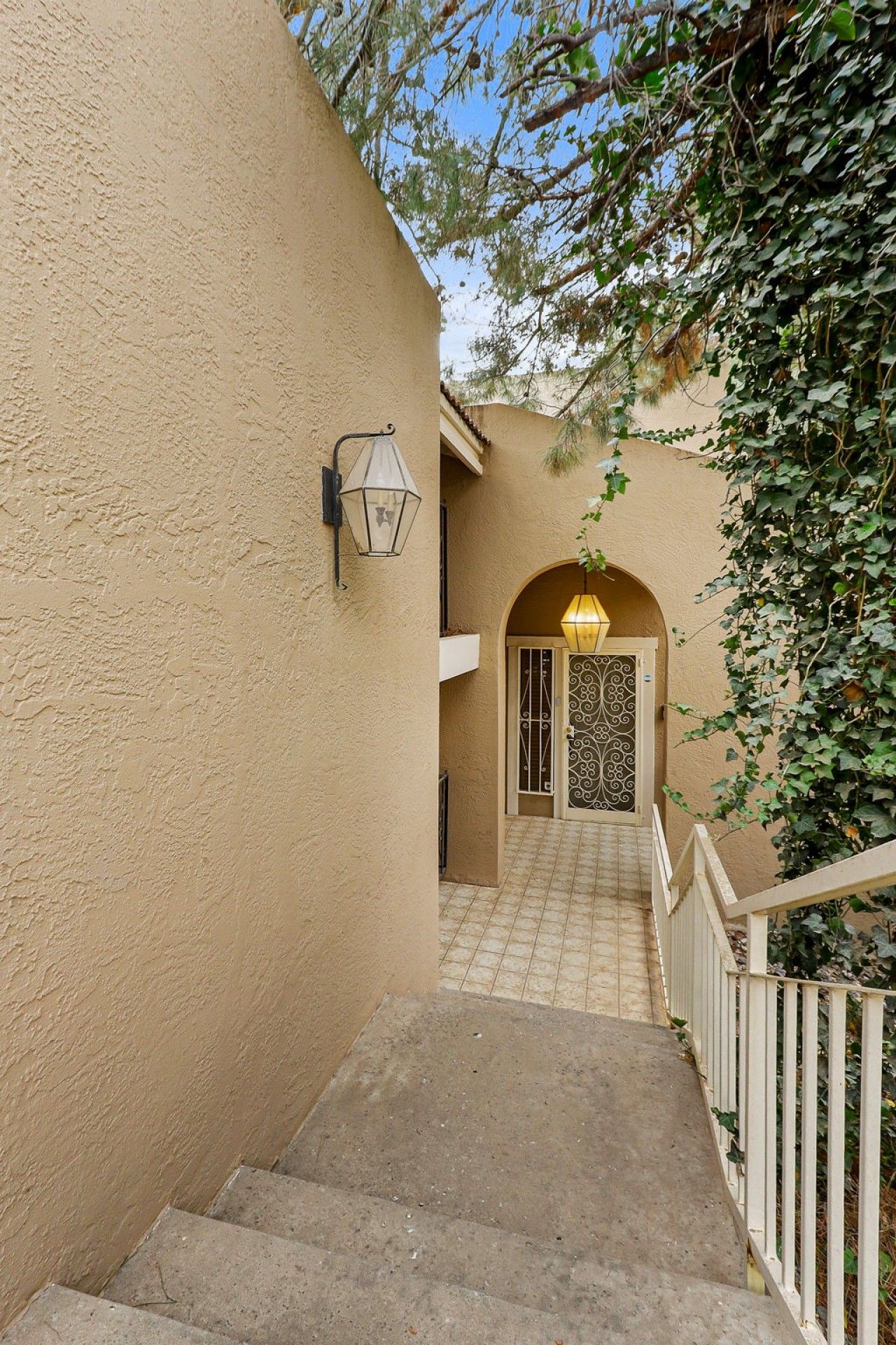 A staircase leading to a door with a decorative gate, a wall-mounted light fixture, and a plant overhanging the stairs.