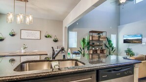 A kitchen with a granite countertop, stainless steel sink, and a dishwasher. There are potted plants on the wall and a built-in shelving unit.