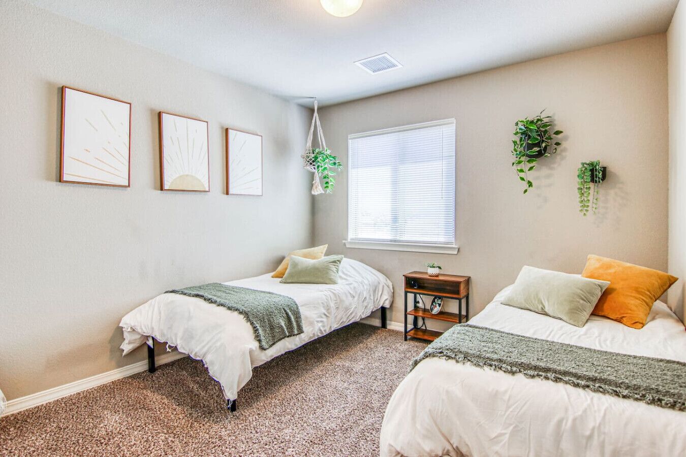 A bedroom with two single beds, a window with blinds, a small bedside table with a plant, and three framed pictures on the wall.