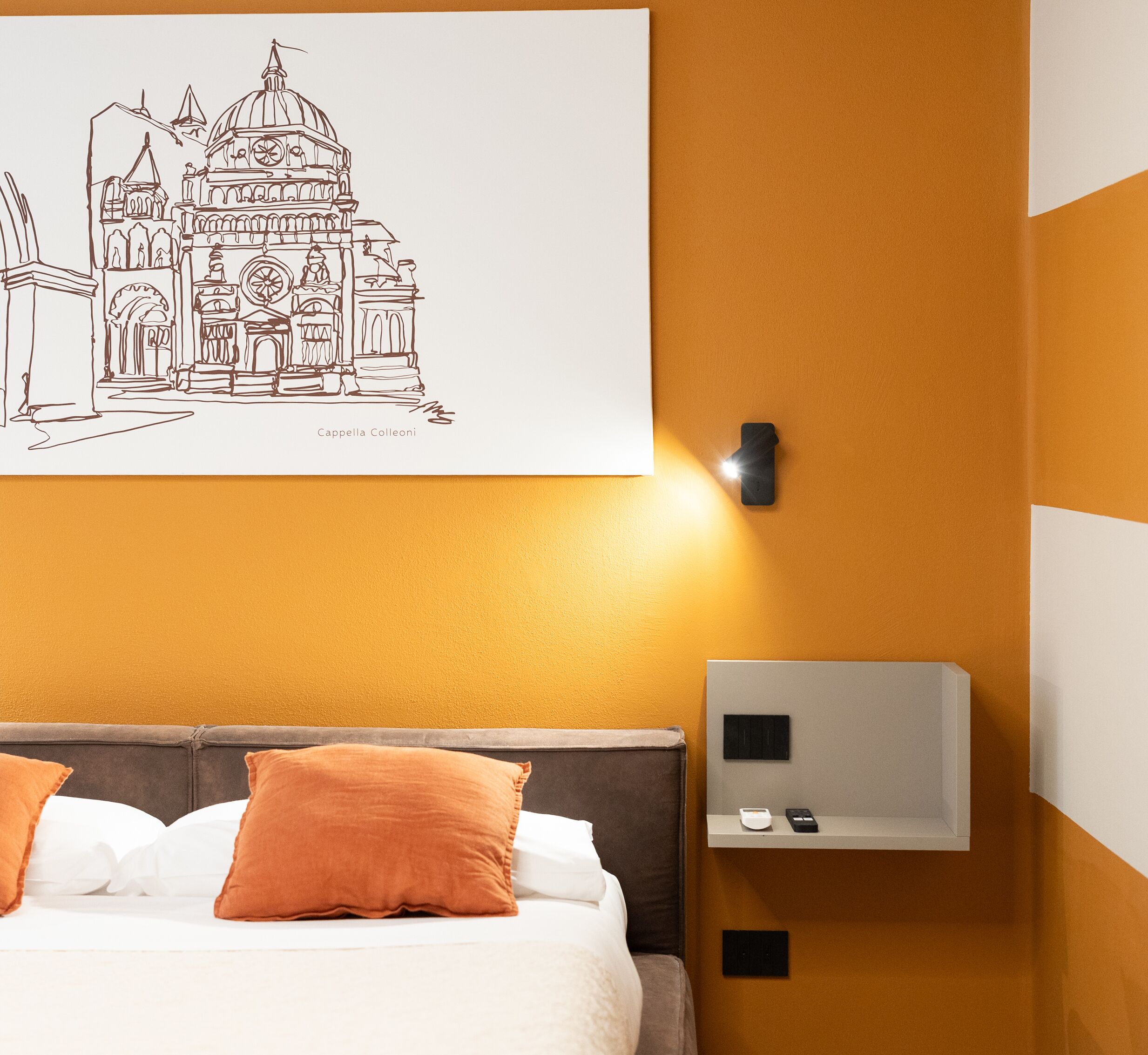 A hotel room with a bed, orange pillows, a wall-mounted light, and a thermostat.