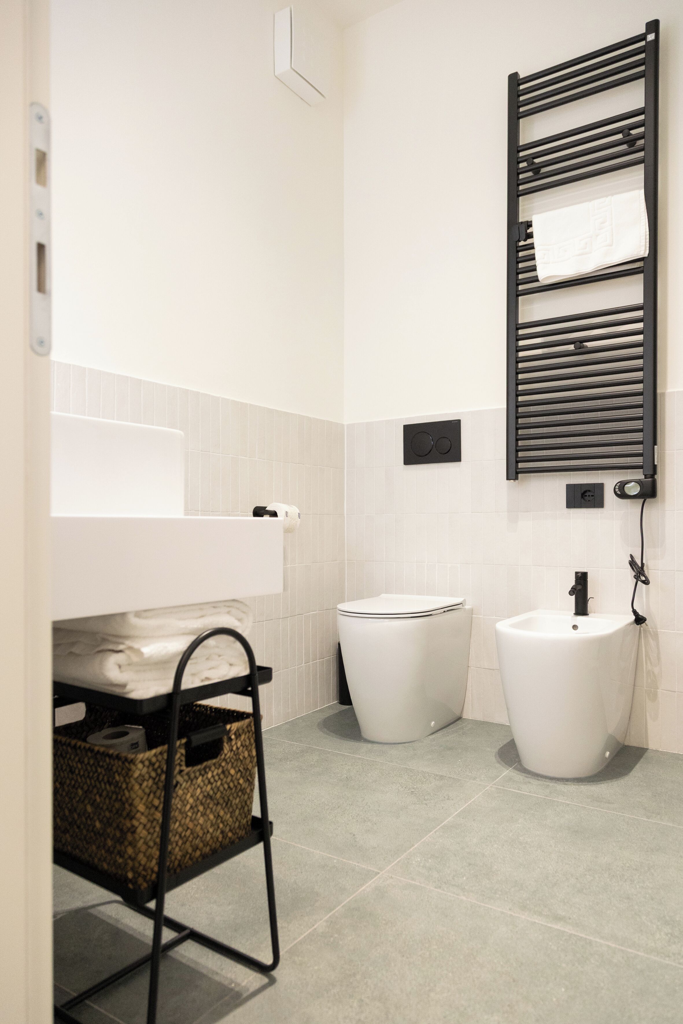 A modern bathroom with a toilet, bidet, and sink. There is a towel rack and a basket under the sink.