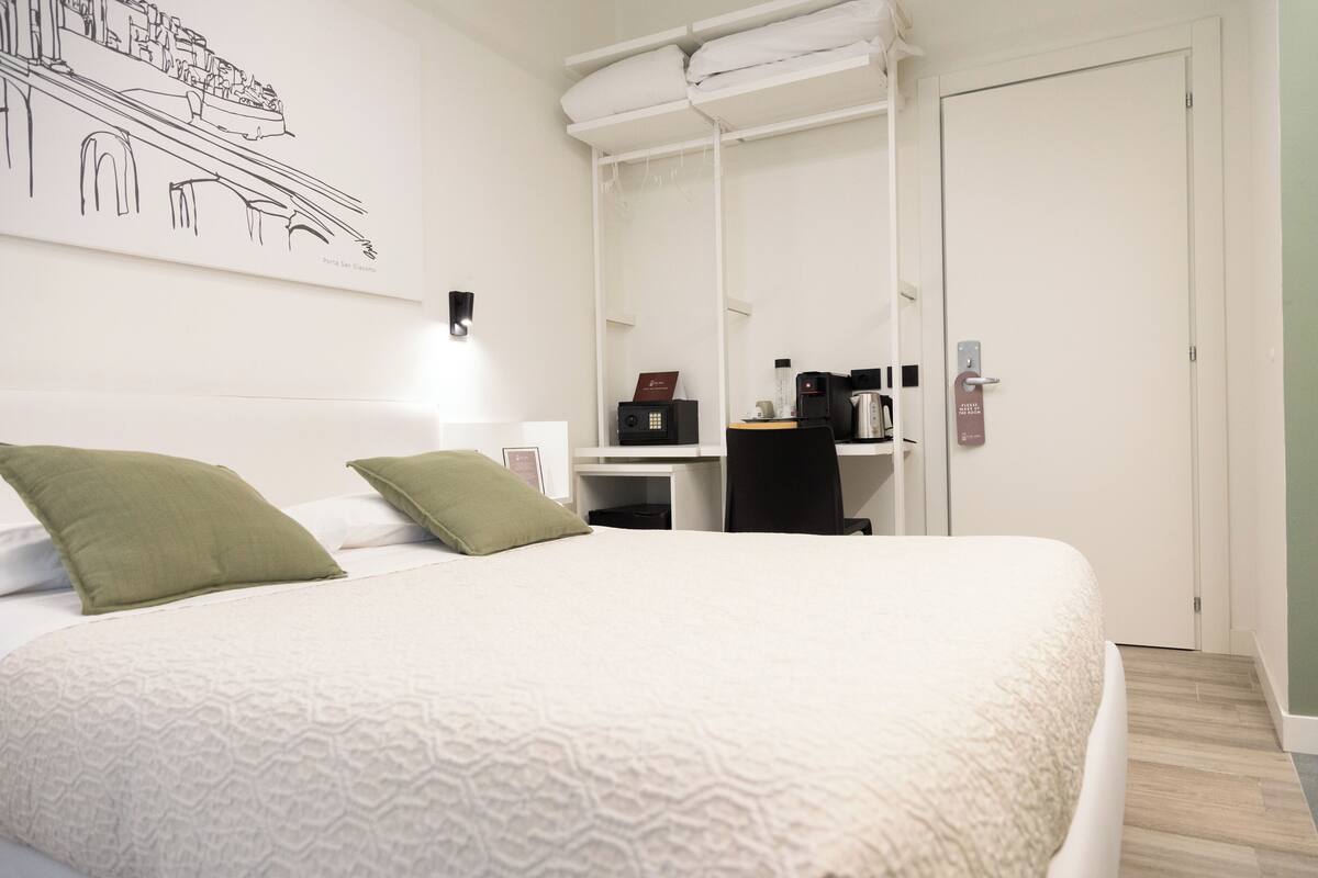 A hotel room with a bed, a desk, a chair, a coffee machine, and a door.
