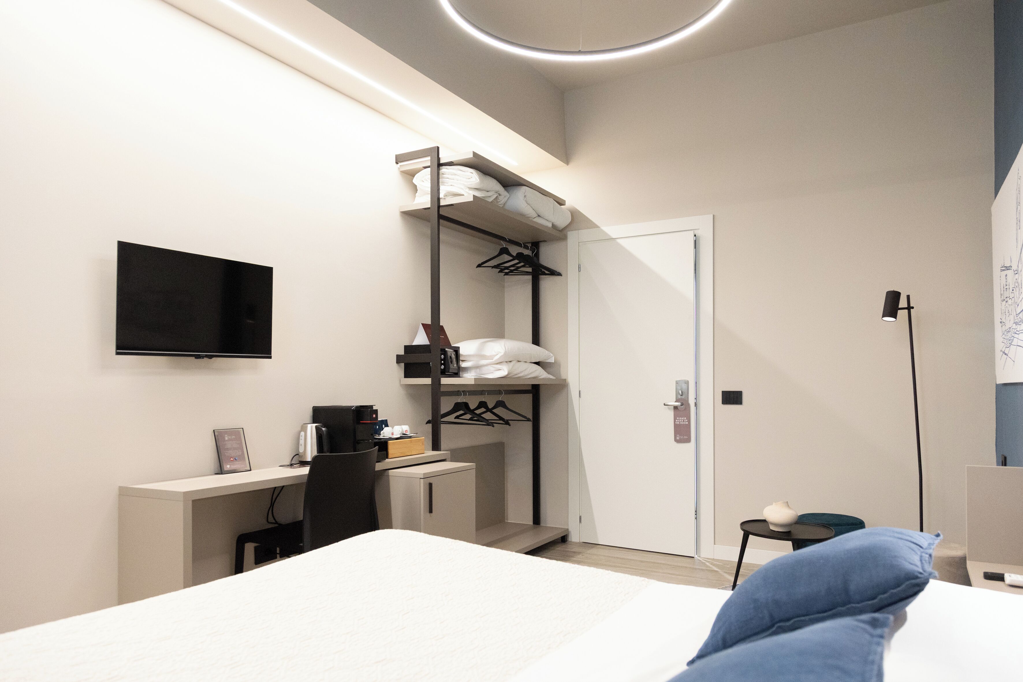 A modern hotel room with a bed, a desk with a computer, a TV mounted on the wall, a closet with hanging clothes, and a small table with a lamp.
