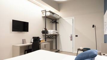 A modern hotel room with a bed, a desk with a computer, a TV mounted on the wall, a closet with hanging clothes, and a small table with a lamp.