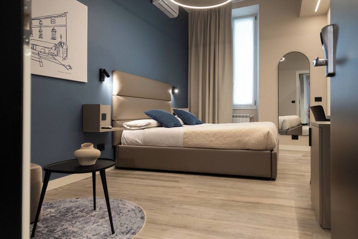 A modern bedroom with a large bed, bedside tables, a small table with a vase, and a wall-mounted mirror.