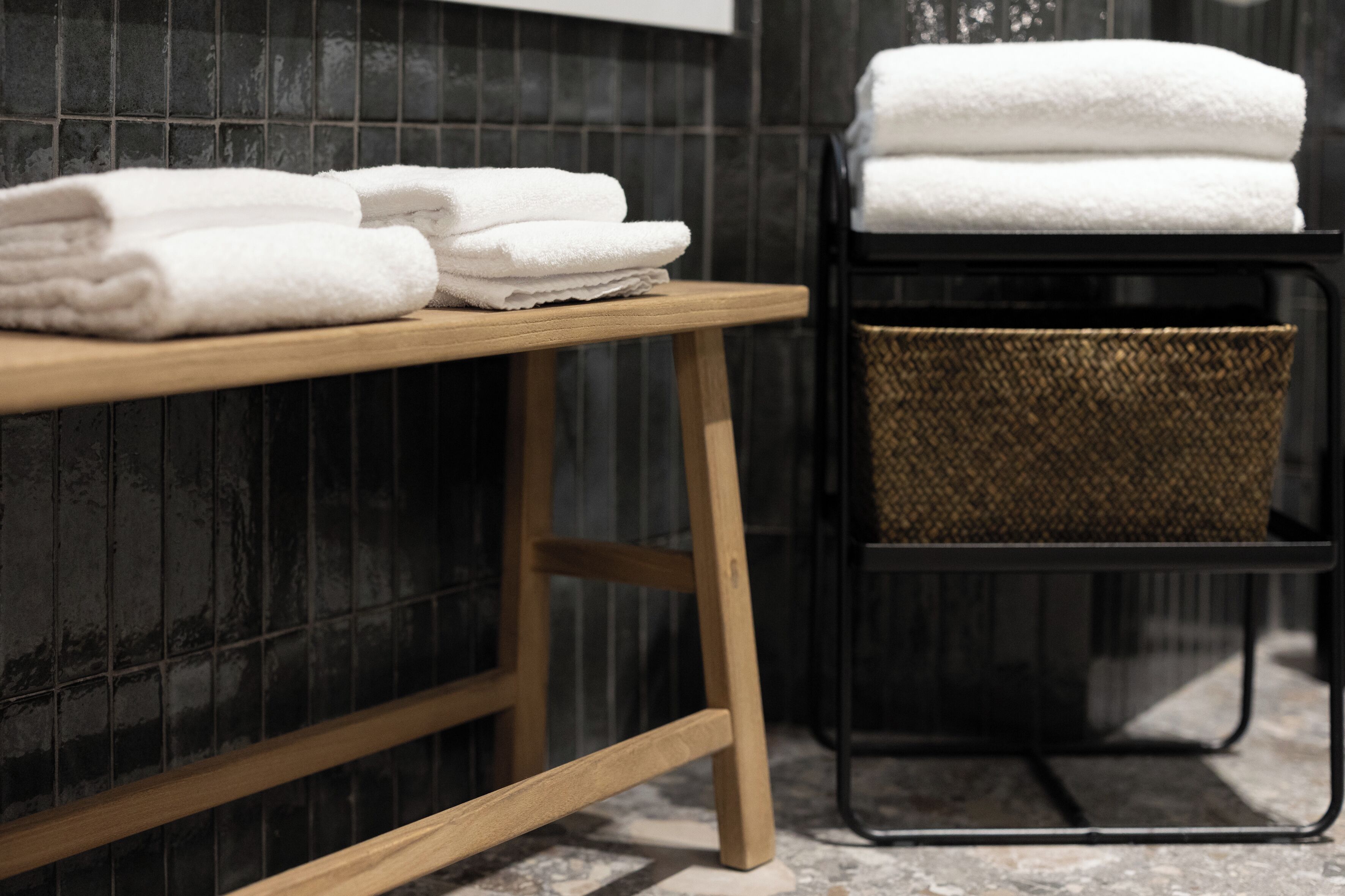 A wooden bench with folded white towels stacked on it and a woven basket on a black metal stand.