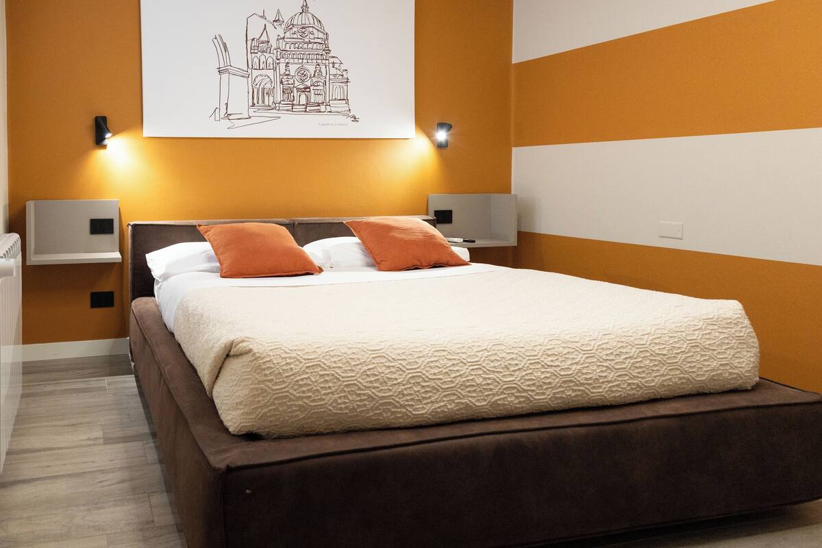 A neatly made bed with a brown headboard, two white pillows, and two orange accent pillows. There are two bedside tables with wall-mounted lamps.