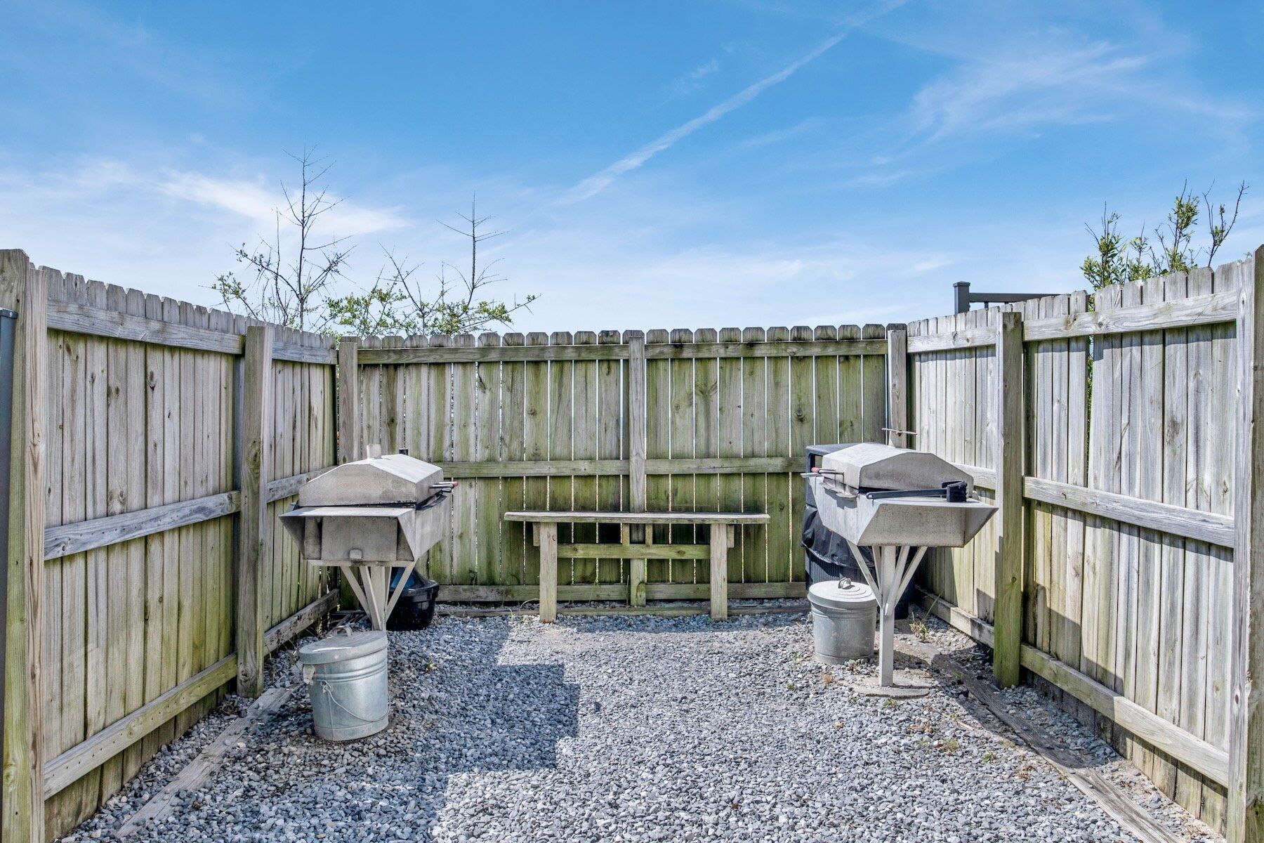 A wooden outdoor barbecue area with two grills and a bench, surrounded by a wooden fence and gravel ground.