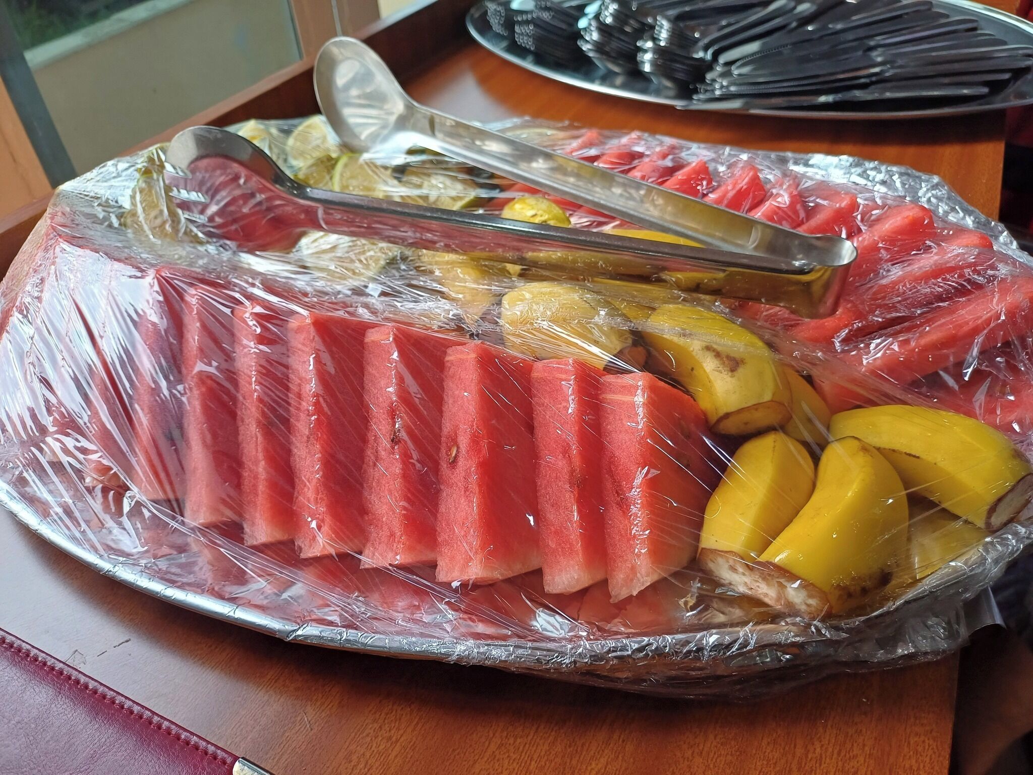 A tray of sliced fruits and vegetables, including watermelon and yellow squash, wrapped in plastic.
