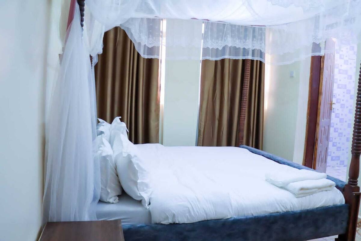 Ein ordentlich bezogenes Bett mit weißen Leinen und einem blauen Kopfteil, ein Nachttisch aus Holz und ein Fenster mit braunen Vorhängen.