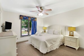 A bedroom with a bed, two bedside tables with lamps, a ceiling fan, a window with curtains, and a television on a stand.