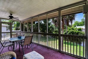 A covered outdoor patio with a table and chairs, a ceiling fan, and a view of a water body and greenery.