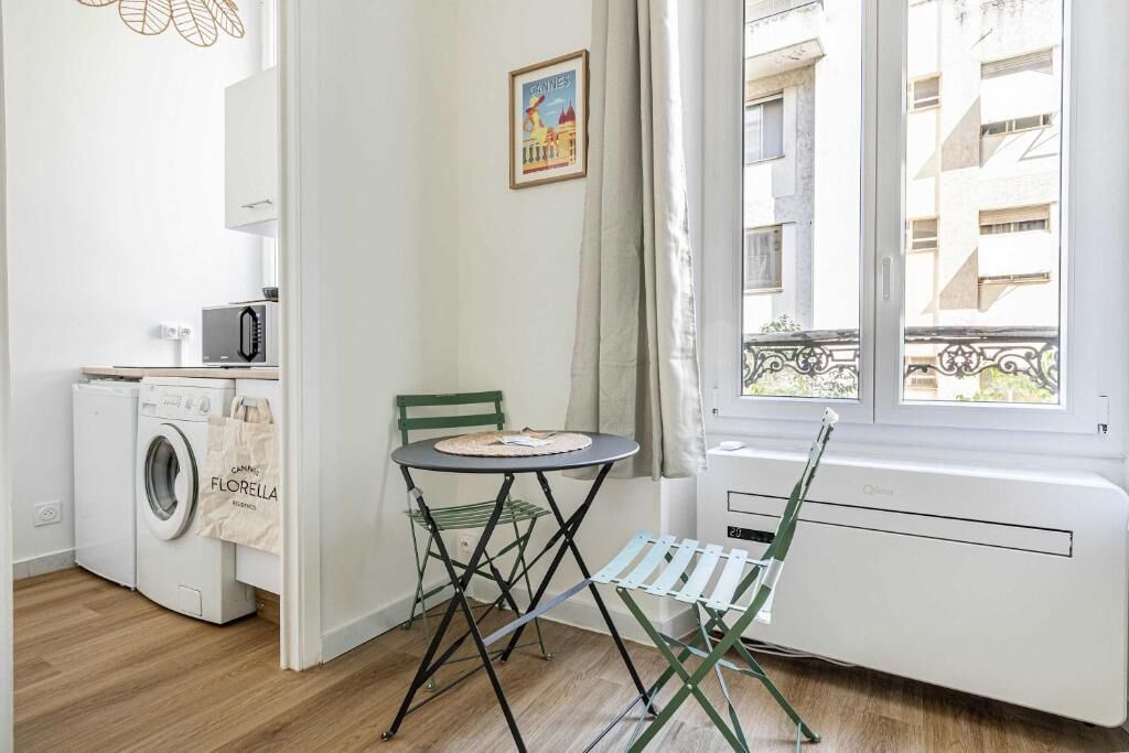 A small, bright room with a washing machine, a table with a chair, and a window with a view of buildings.
