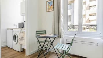 A small, bright room with a washing machine, a table with a chair, and a window with a view of buildings.