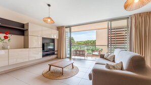 Ein modernes Wohnzimmer mit Flachbildfernseher, einer Couch, einem Couchtisch und einem Balkon mit Blick auf Palmen.