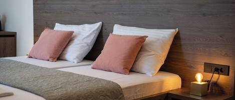 Una cama doble con cabecera de madera, dos almohadas, una mesita de noche con lámpara y otra mesita auxiliar.