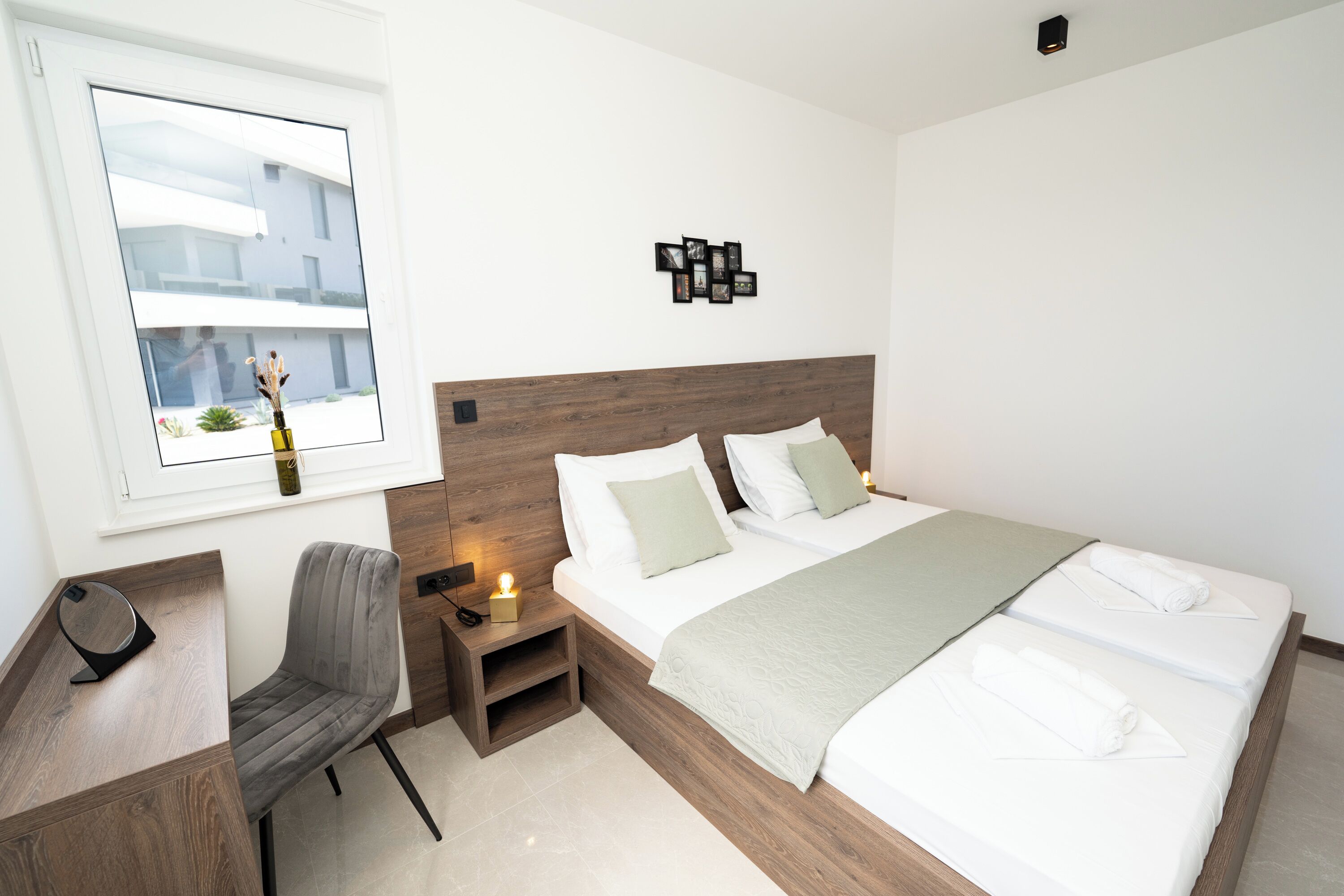A modern hotel room with a wooden headboard, a desk with a chair, a bedside table with a lamp, and a view of buildings outside the window.