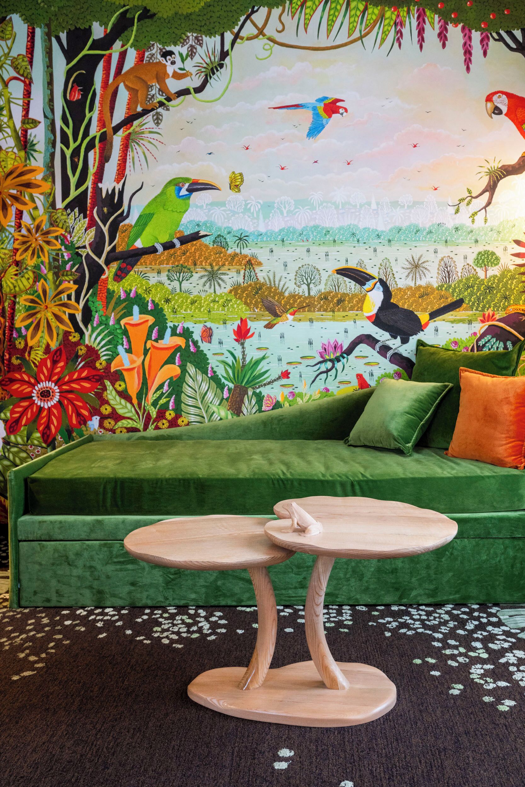 Ein grünes Sofa mit einem runden Holztisch davor, ein lebhaftes Wandgemälde mit Vögeln und Blattwerk, und ein gemustertes Teppich.