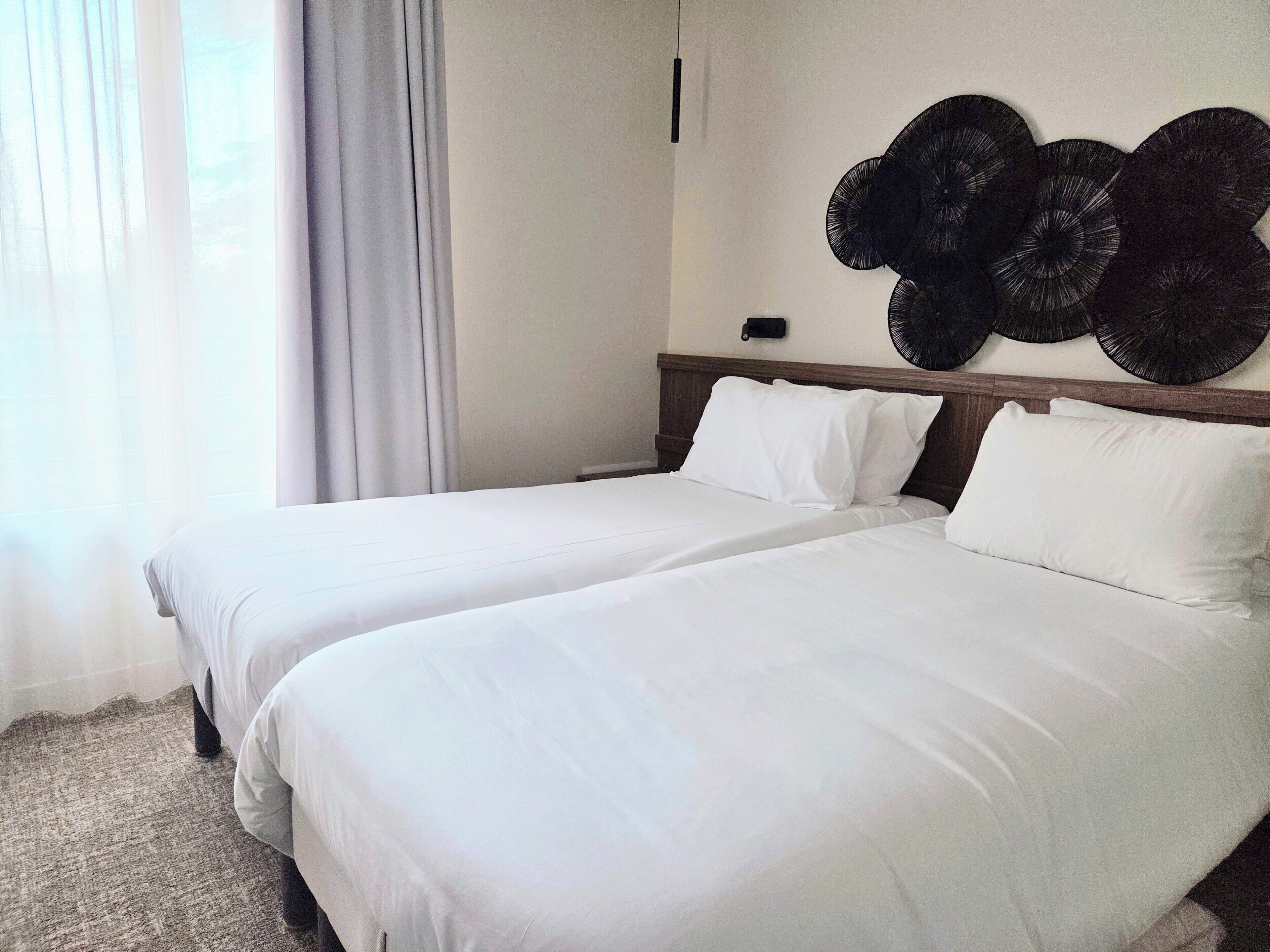 A hotel room with two beds, a large window with sheer curtains, and a wall-mounted decorative item.