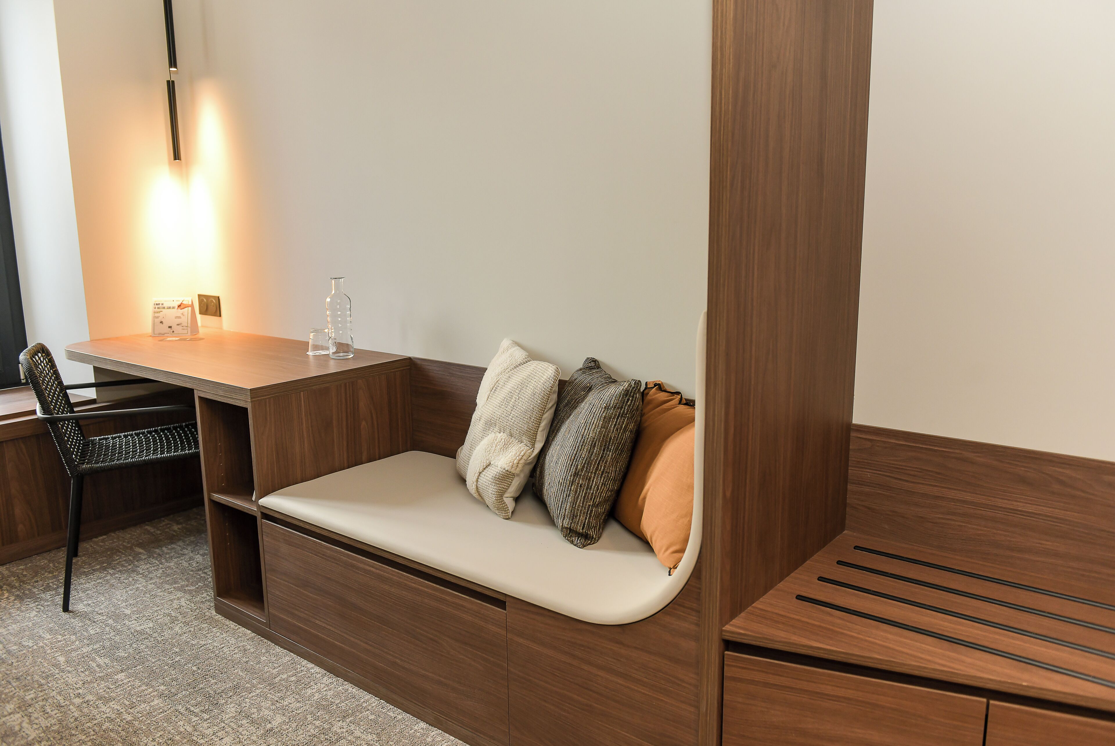 A modern, compact room with a built-in desk, a cushioned bench with pillows, a chair, and a lamp.
