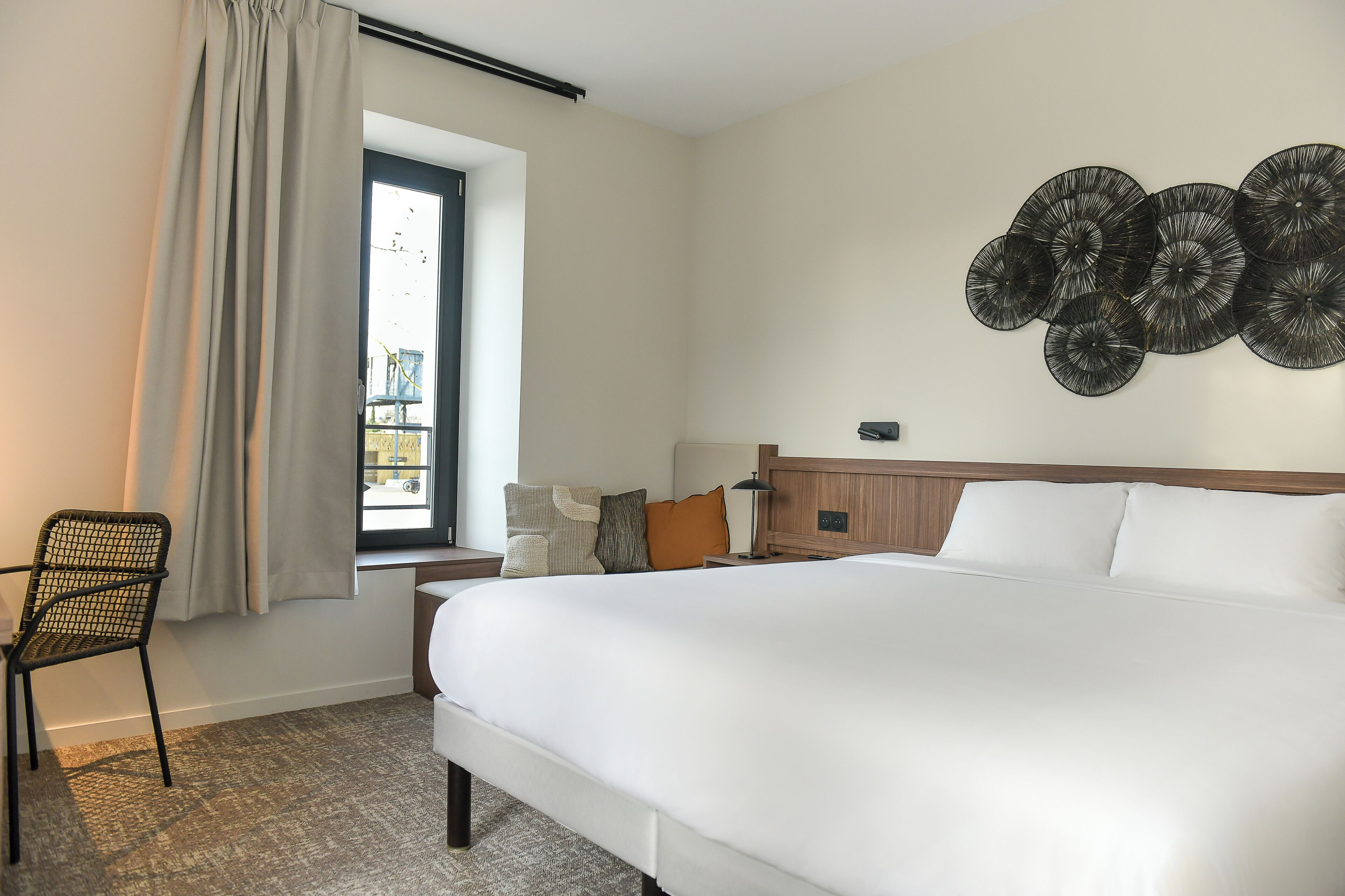 A hotel room with a large bed, a window with curtains, a wall-mounted art piece, a chair, and a bedside table with a lamp.